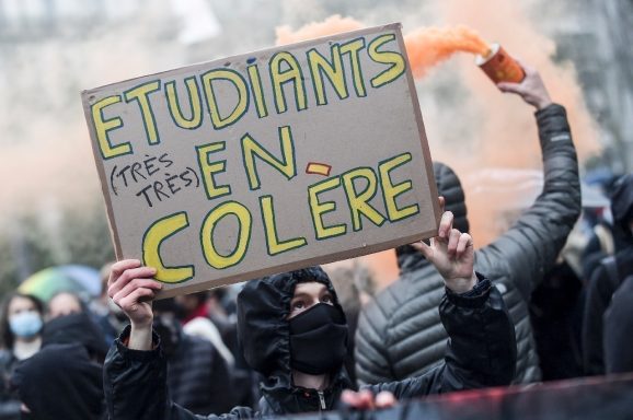 Un manifestant tient une pancarte : "Étudiants en colère", entouré de fumée orange.